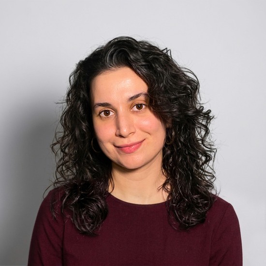 Portrait of a woman with long curly dark hair and a slight smile wearing a maroon shirt against a gray background.